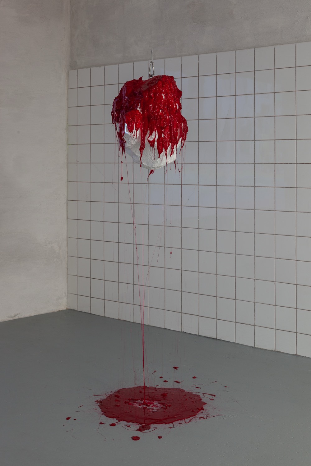 Varda by Diana Burkot, 2025: a white stone form hanging in the corner of the tiled room, red substance running from it into a pool on the floor